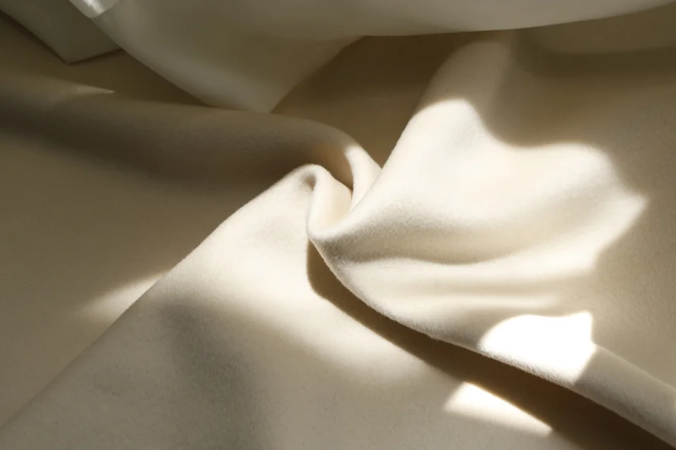 Ivory-coloured coating fabric arranged in broad folds under directional light.